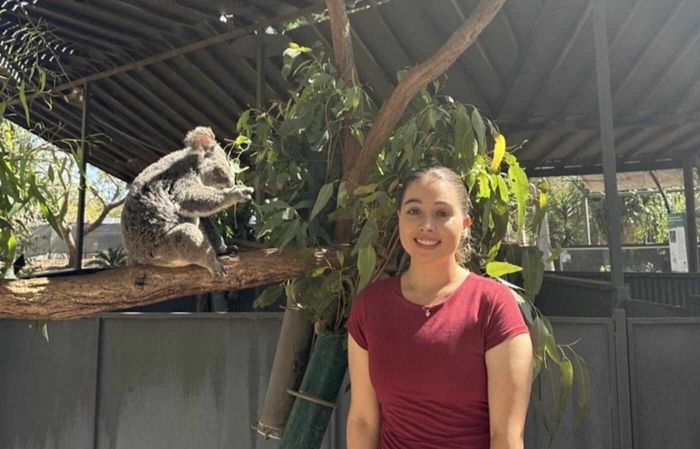 Anna Benson standing next to a tree with a koala eating leaves to the left of the photo.