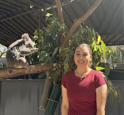 Anna Benson standing next to a tree with a koala eating leaves to the left of the photo.